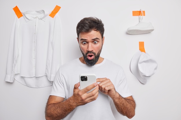 Homem barbudo chocado olha para o telefone na tela e vende roupas desnecessárias online olha para os olhos arregalados usa camiseta casual isolada na parede branca lê notícias chocantes