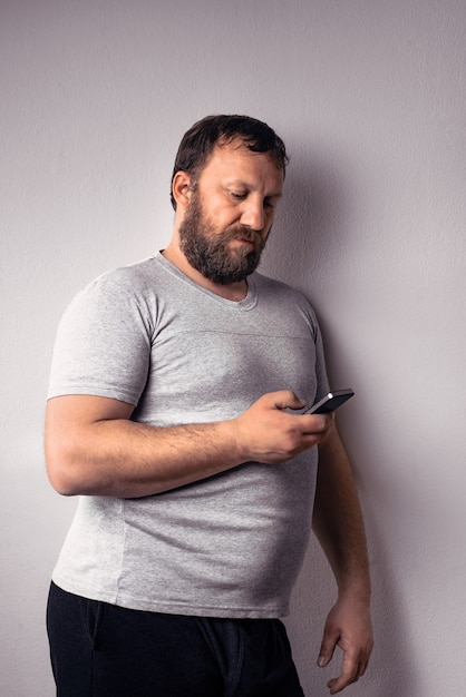 Homem barbudo com camiseta cinza segurando o celular e encostado na parede cinza