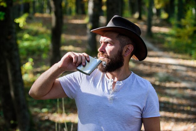 Homem barbudo com chapéu de cowboy anda no parque ao ar livre homem hipster relaxa na floresta de outono Tempo de primavera acampar e caminhadas macho maduro com olhar brutal beber álcool de frasco metálico Cowboy bonito