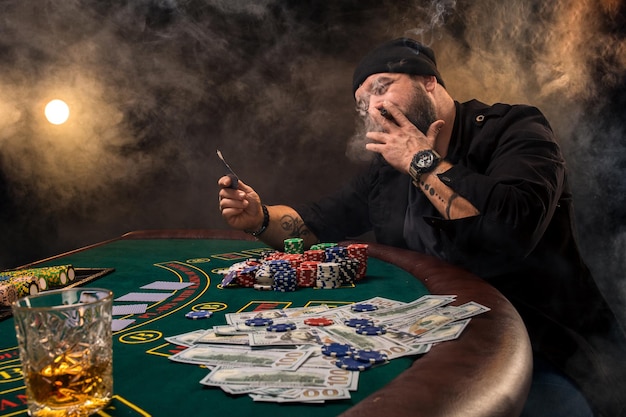 Homem barbudo com charuto e vidro, sentado à mesa de pôquer em um cassino. Jogos de azar, cartas de jogar e roleta.