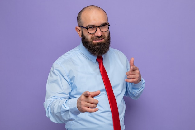Homem barbudo com gravata vermelha e camisa azul usando óculos, olhando para a câmera, feliz e positivo, sorrindo apontando com o dedo indicador para a câmera em pé sobre o fundo roxo
