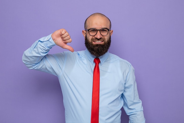 Homem barbudo com gravata vermelha e camisa azul usando óculos, olhando para a câmera. Sorriso feliz e positivo, apontando alegremente para si mesmo em pé sobre um fundo roxo