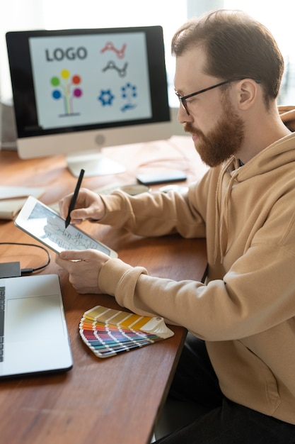 Homem barbudo concentrado com capuz sentado à mesa de madeira e usando um tablet enquanto trabalha no design da marca