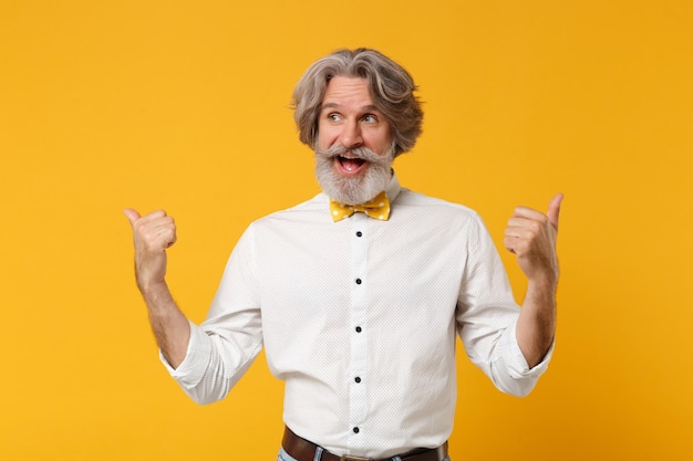 Homem barbudo de bigode grisalho idoso alegre em gravata borboleta de camisa branca posando isolado em fundo amarelo laranja no estúdio. Conceito de estilo de vida de pessoas. Mock up espaço de cópia. Apontando os polegares de lado.