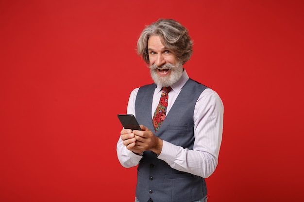 Homem barbudo de bigode grisalho idoso alegre em gravata colorida de colete de camisa clássica posando isolado em fundo vermelho no estúdio. Conceito de estilo de vida de pessoas. Mock up espaço de cópia. Segurando o celular.