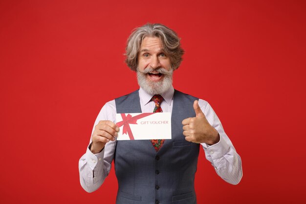 Homem barbudo de bigode grisalho idoso alegre na gravata colorida clássica do colete da camisa isolada no fundo vermelho. conceito de estilo de vida de pessoas. mock up espaço de cópia. segure o vale-presente mostrando o polegar para cima.