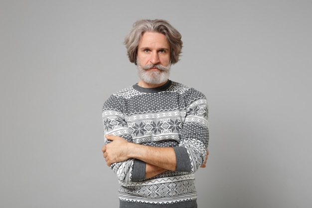 Homem barbudo de bigode grisalho idoso bonito na camisola de malha posando isolado no fundo da parede cinza, retrato de estúdio. Conceito de estilo de vida de pessoas. Mock up espaço de cópia. De mãos dadas cruzadas.