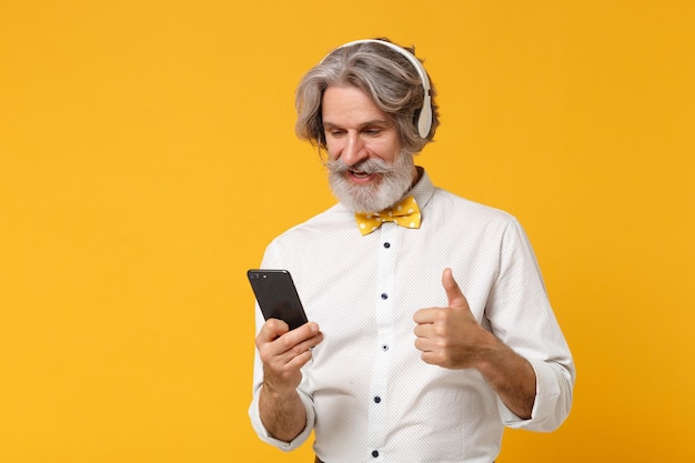 Homem barbudo de bigode grisalho idoso em gravata borboleta de camisa isolada em fundo amarelo. Conceito de estilo de vida de pessoas. Mock up espaço de cópia. Ouça música com fones de ouvido segure o celular, mostrando o polegar para cima.