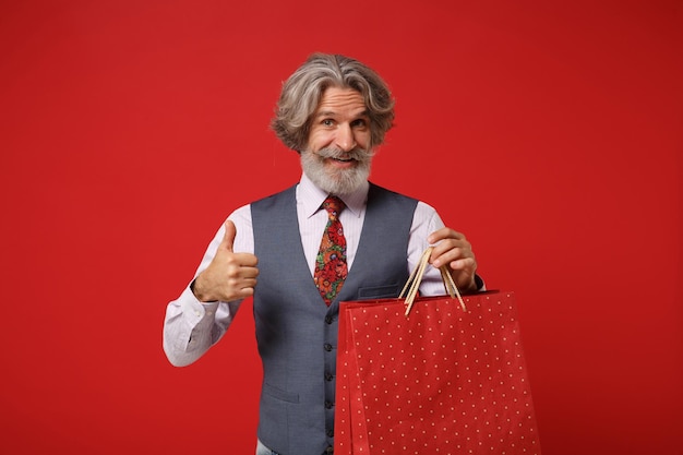 Homem barbudo de bigode grisalho idoso em gravata de colete de camisa isolada em fundo vermelho. Conceito de estilo de vida de pessoas. Mock up espaço de cópia. Segure o saco do pacote com a compra após as compras, mostrando o polegar para cima.