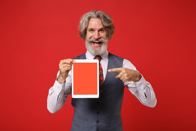 Foto homem barbudo de bigode grisalho idoso em gravata de colete de camisa posando isolado em fundo vermelho. conceito de estilo de vida de pessoas. mock up espaço de cópia. apontando o dedo no computador tablet com tela vazia em branco.