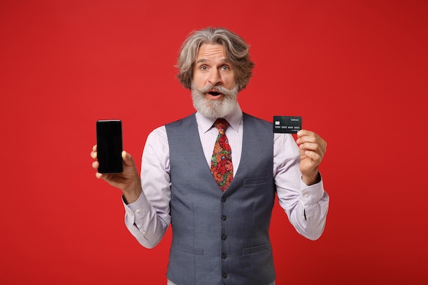 Homem barbudo de bigode grisalho idoso em gravata de colete de camisa posando isolado em fundo vermelho. Conceito de estilo de vida de pessoas. Mock up espaço de cópia. Segure o celular com tela vazia em branco, cartão de banco de crédito.