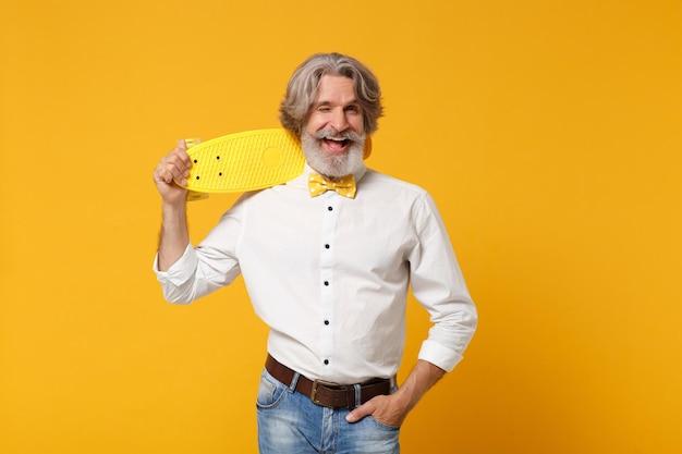 Foto homem barbudo de bigode grisalho idoso engraçado na camisa branca e gravata borboleta isolada no retrato de estúdio de fundo laranja amarelo. conceito de estilo de vida de pessoas. mock up espaço de cópia. segure o skate, piscando.