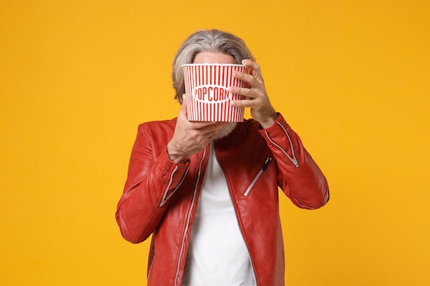 Foto homem barbudo de bigode grisalho idoso na jaqueta de couro vermelha isolada no fundo da parede amarelo laranja. emoções de pessoas no conceito de cinema. assistindo filme, cobrindo o rosto com balde de pipoca.