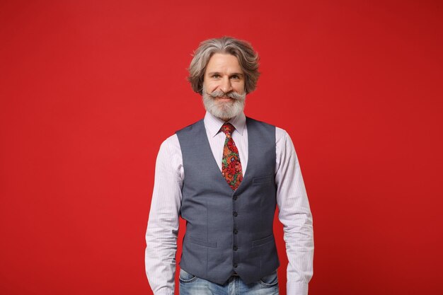 Homem barbudo de bigode grisalho idoso sorridente na camisa clássica, colete, gravata colorida posando isolado no retrato de estúdio de fundo vermelho. conceito de estilo de vida de emoções sinceras de pessoas. mock up espaço de cópia.