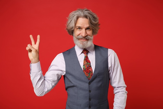 Homem barbudo de bigode grisalho idoso sorridente no colete de camisa clássico e gravata colorida posando isolado em fundo vermelho no estúdio. Conceito de estilo de vida de pessoas. Mock up espaço de cópia. Mostrando sinal de vitória.