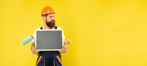 Homem barbudo de capacete e roupas de trabalho segura pincel de rolo de pintura e quadro-negro com espaço de cópia