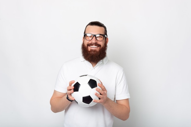 Homem barbudo de óculos está segurando uma bola de futebol e sorrindo para a câmera.