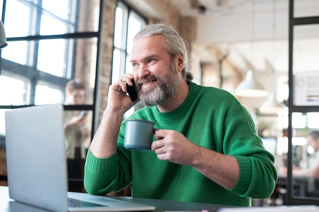 Homem barbudo de suéter casual tomando café e falando ao telefone enquanto está sentado à mesa com o laptop no escritório