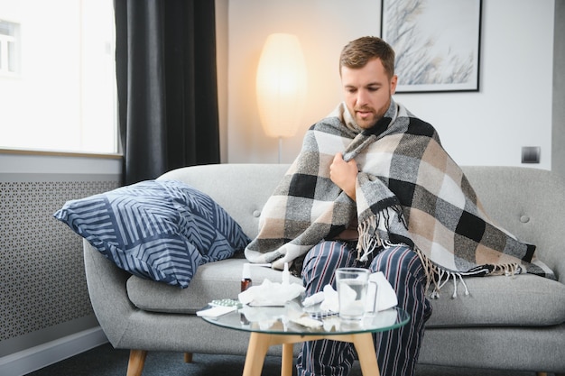 Homem barbudo doente que tem resfriado ou gripe sazonal sentado no sofá em casa Cara com febre vestindo manta quente tremendo com expressão de rosto preocupado
