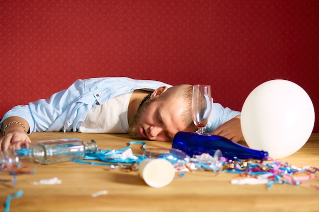 Homem barbudo dormindo à mesa em um quarto bagunçado depois da despedida de solteiro, homem cansado depois da festa em casa