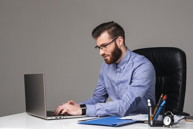Homem barbudo e elegante de óculos, satisfeito, usando um laptop enquanto está sentado à mesa no escritório