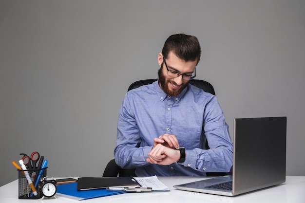 Homem barbudo e elegante de óculos, usando seu relógio de pulso enquanto está sentado à mesa no escritório, sorrindo