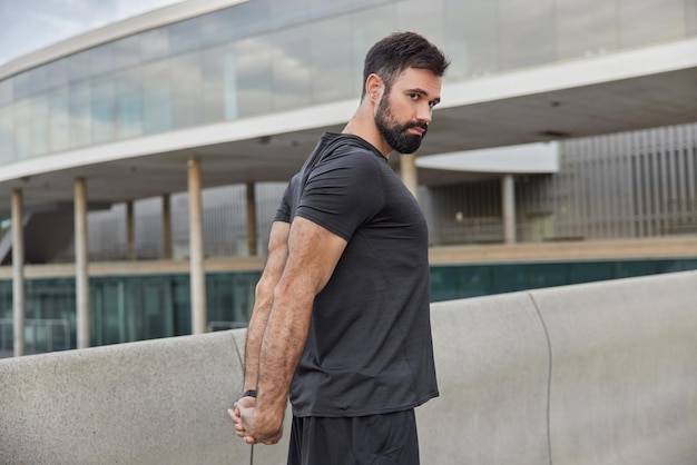 Homem barbudo e esportivo estende os braços musculosos, vestido com uma camiseta preta, aquece antes que o treino concentrado na distância tenha suportes de treinamento regulares contra o meio urbano. exercícios de atleta masculino