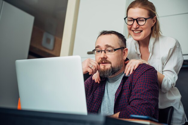 Foto homem barbudo e mulher com óculos usando laptop no interior de casa, se comunicar com amigos online, rir e fazer careta.