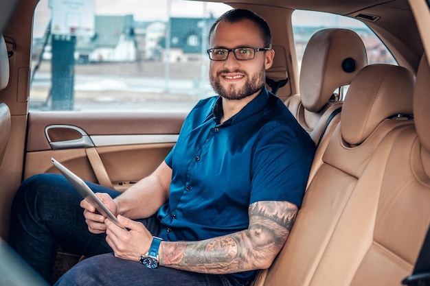 Homem barbudo elegante em óculos com tatuagem no braço usando tablet PC portátil no banco traseiro de um carro.