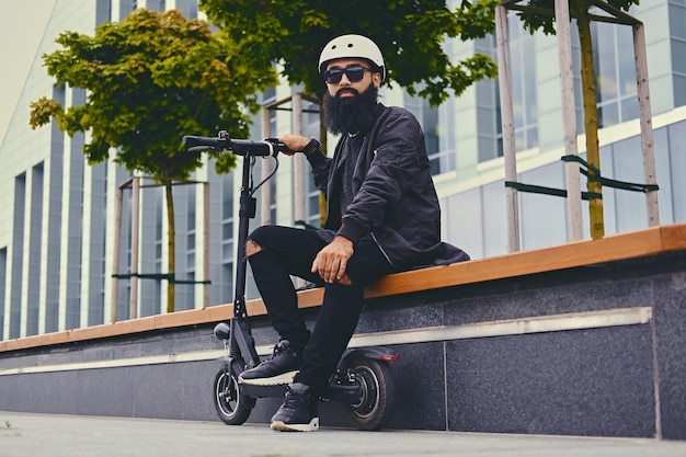 Homem barbudo elegante em óculos de sol relaxando em um banco depois de andar de scooter elétrico no centro da cidade.