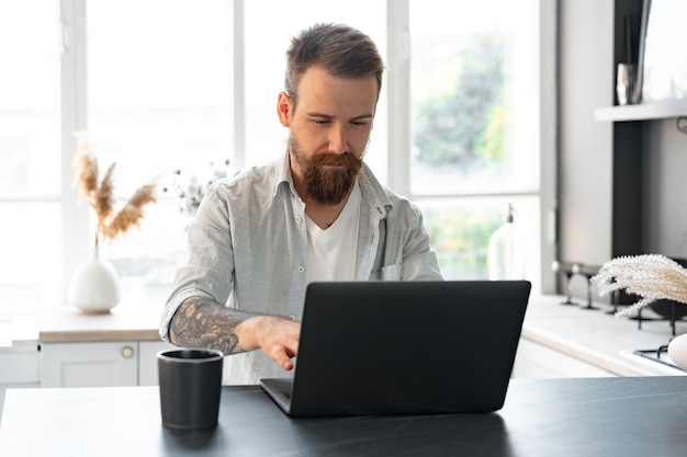 Homem barbudo elegante trabalhando no laptop em casa sentado à mesa