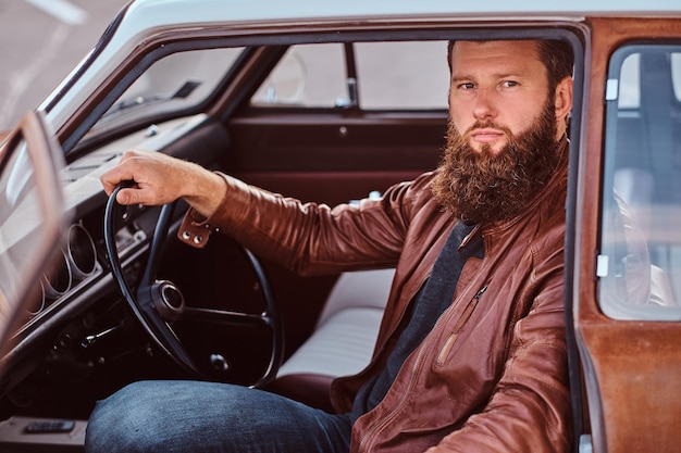 Homem barbudo elegante, vestido com jaqueta de couro marrom, senta-se ao volante de um carro retrô sintonizado com porta aberta.