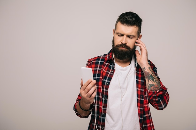 Homem barbudo em camisa quadriculada ouvindo música com fones de ouvido e smartphone isolado em cinza