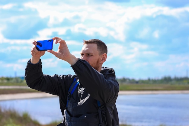 Homem barbudo em capa de chuva preta em um fundo de paisagem de mar azul com smartphone nas mãos tira fotografia selfie
