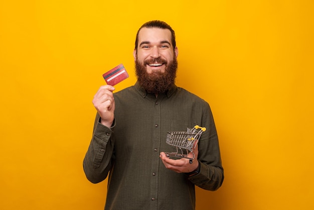 Homem barbudo em êxtase está segurando um pequeno carrinho de compras e um cartão de crédito studio atirou sobre fundo amarelo