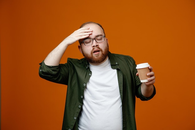 Homem barbudo em roupas casuais usando óculos segurando a xícara de café parecendo confuso segurando a mão na testa por erro em pé sobre fundo laranja