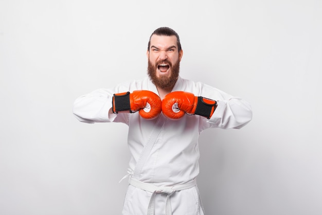 Homem barbudo empolgado e espantado em taekwondo dobok comemorando sobre uma parede branca