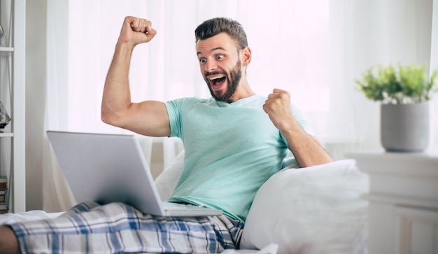 Homem barbudo empolgado e feliz fã da vitória com um laptop em uma grande cama branca em casa gritando durante a celebração