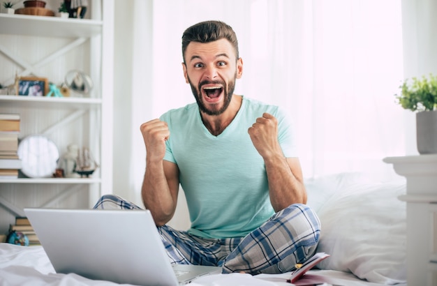 Homem barbudo empolgado e feliz fã da vitória com um laptop em uma grande cama branca em casa gritando durante a celebração