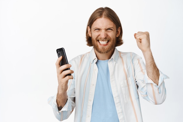 Homem barbudo empolgado ganhando no celular, segurando o smartphone e sorrindo para a alegria, ganhando dinheiro com o app para smartphone, fazendo compras online, parado no branco