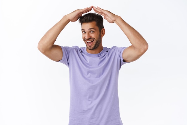 Homem barbudo encantador alegre em camiseta roxa elegante levanta as mãos acima da cabeça e fazendo gesto de telhado ou casa sorrindo divertido não tem nada se preocupar como se proteger fundo branco