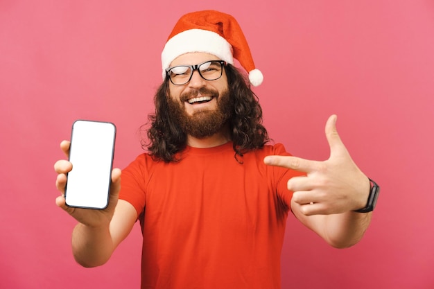 Homem barbudo encaracolado usando um boné de Natal aponta para a tela vazia do telefone