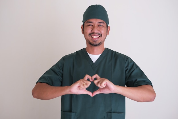 Homem barbudo enfermeira asiática sorrindo amigavelmente e dando forma de coração de amor usando os dedos