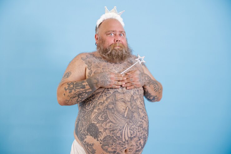Foto homem barbudo engraçado com excesso de peso usando coroa de brinquedo cobrindo os seios em fundo azul claro