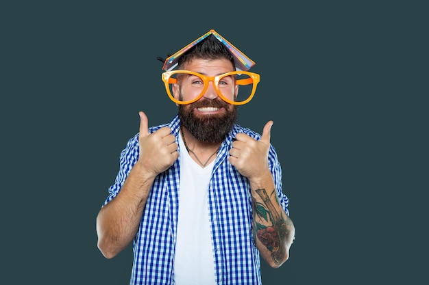 Homem barbudo engraçado em óculos de festa com livro na cabeça polegar para cima