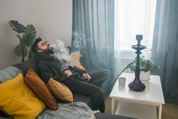 Homem barbudo está fumando cachimbo de água em casa e soprando nuvem de fumaça, tempo frio e conceito de descanso