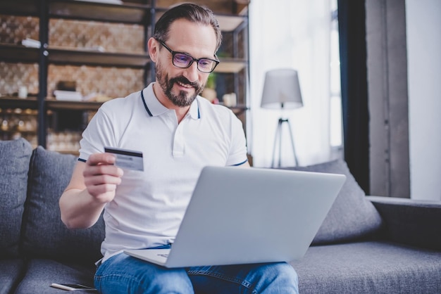Homem barbudo fazendo compras online com cartão de crédito e usando laptop em casa