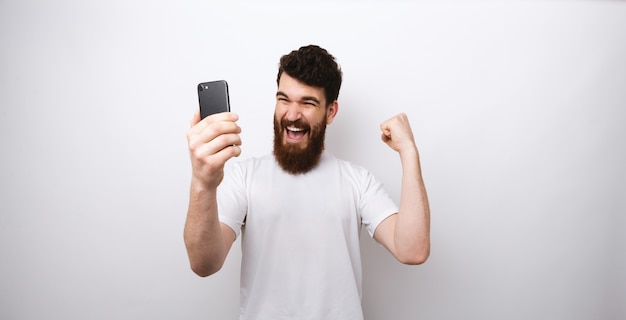 Homem barbudo fazendo um vencedor gesto e olhando para o celular em fundo branco.