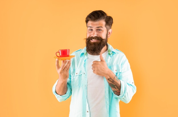 homem barbudo feliz beber café da manhã mostrando o polegar para cima em um copo de café de fundo amarelo