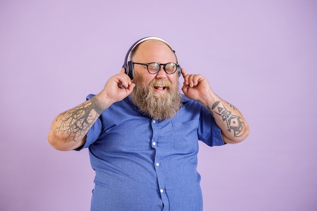 Homem barbudo feliz com excesso de peso ouve música com fones de ouvido no fundo roxo